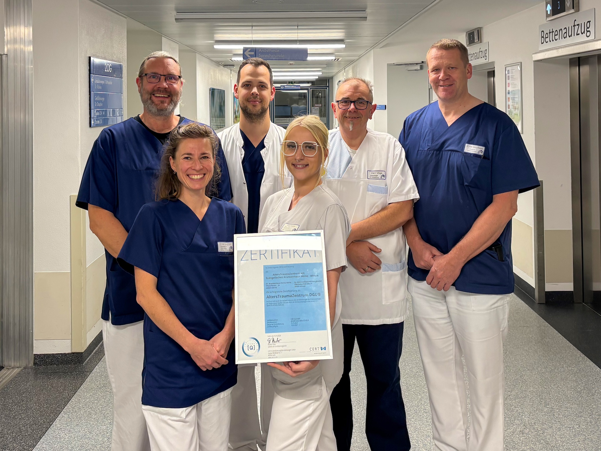 Sechs lächelnde Mitarbeiter des Gesundheitswesens stehen in einem Flur; einer hält eine gerahmte Urkunde.