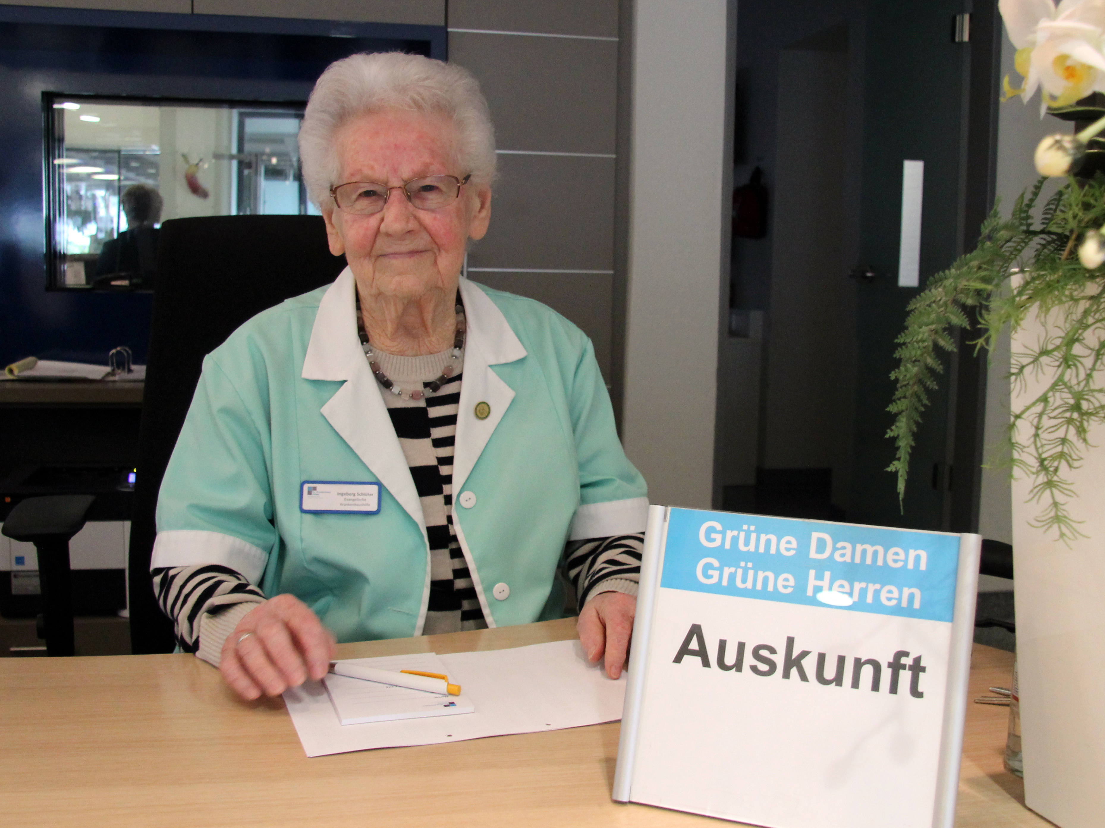 Eine ältere Frau in grüner Uniform sitzt an einem Informationsschalter und hält ein Schild mit der Aufschrift "Auskunft" und einen Stift in der Hand.