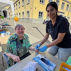 Eine junge Frau mit dunklen Haaren und blauen Handschuhen steht vor einer Frau, die auf einem Stuhl sitzt. Sie hält ihren Finger.