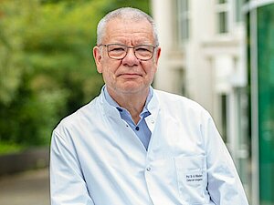 Mann mit wenig Haaren und Brille steht draußen vor einem Gebäude. Er trägt einen weißen Kittel.