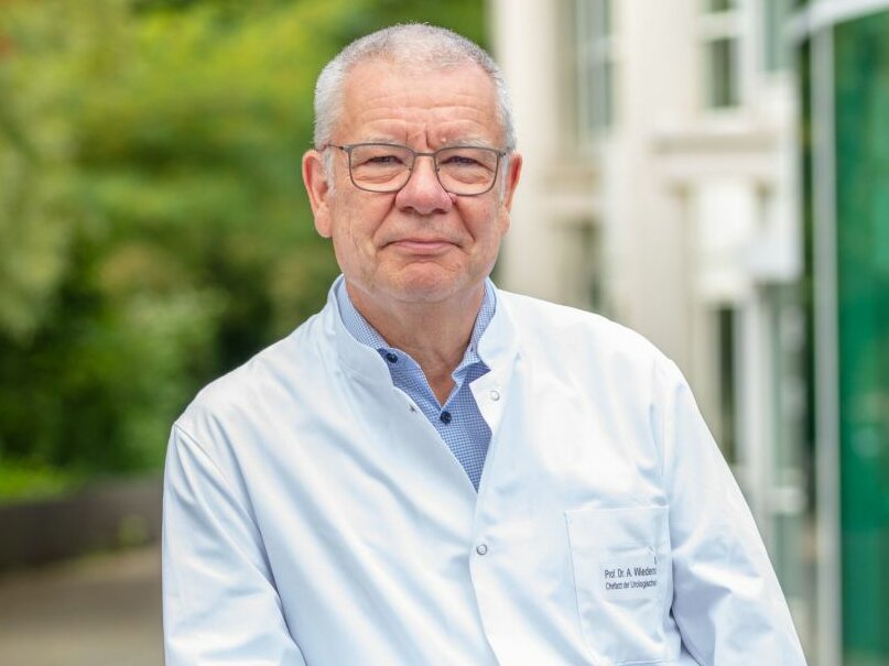 Mann mit wenig Haaren und Brille steht draußen vor einem Gebäude. Er trägt einen weißen Kittel.