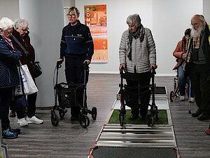 Ältere Menschen mit Rollatoren navigieren auf einem simulierten unebenen Gehweg, beobachtet von einer kleinen Gruppe in einem Innenraum.