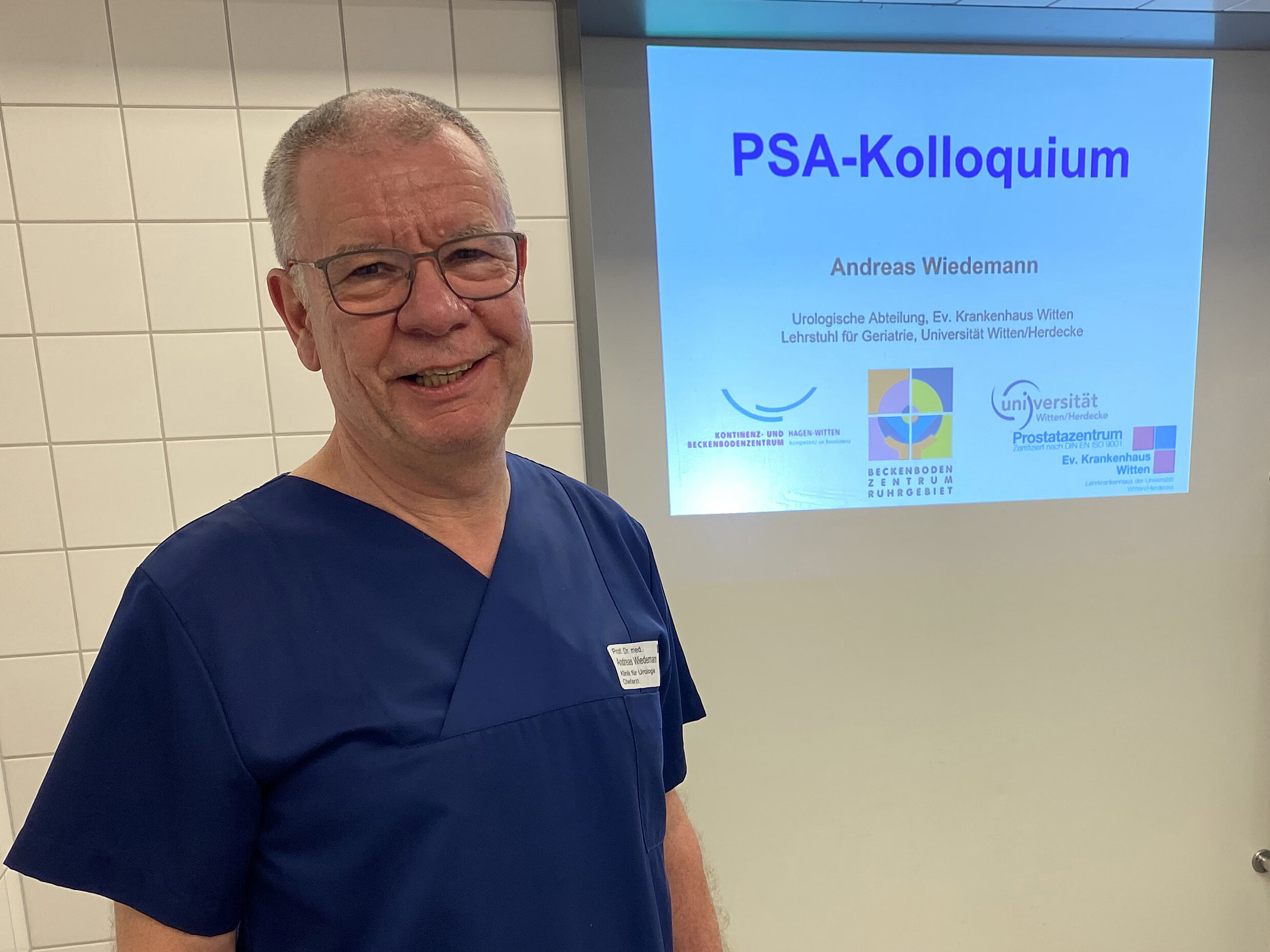Ein lächelnder Mann in blauem Kittel steht vor einem Bildschirm mit der Aufschrift "PSA-Kolloquium" in einem gekachelten Raum.