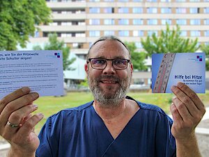 Ein Mann mit Brille und Zopf steht vor einem Krankenhaus. Er hält zwei Karten in den Händen. Darauf steht "Hilfe bei Hitze" und "5 Tipps, wie Sie den heißen Tagen die kalte Schulter zeigen".