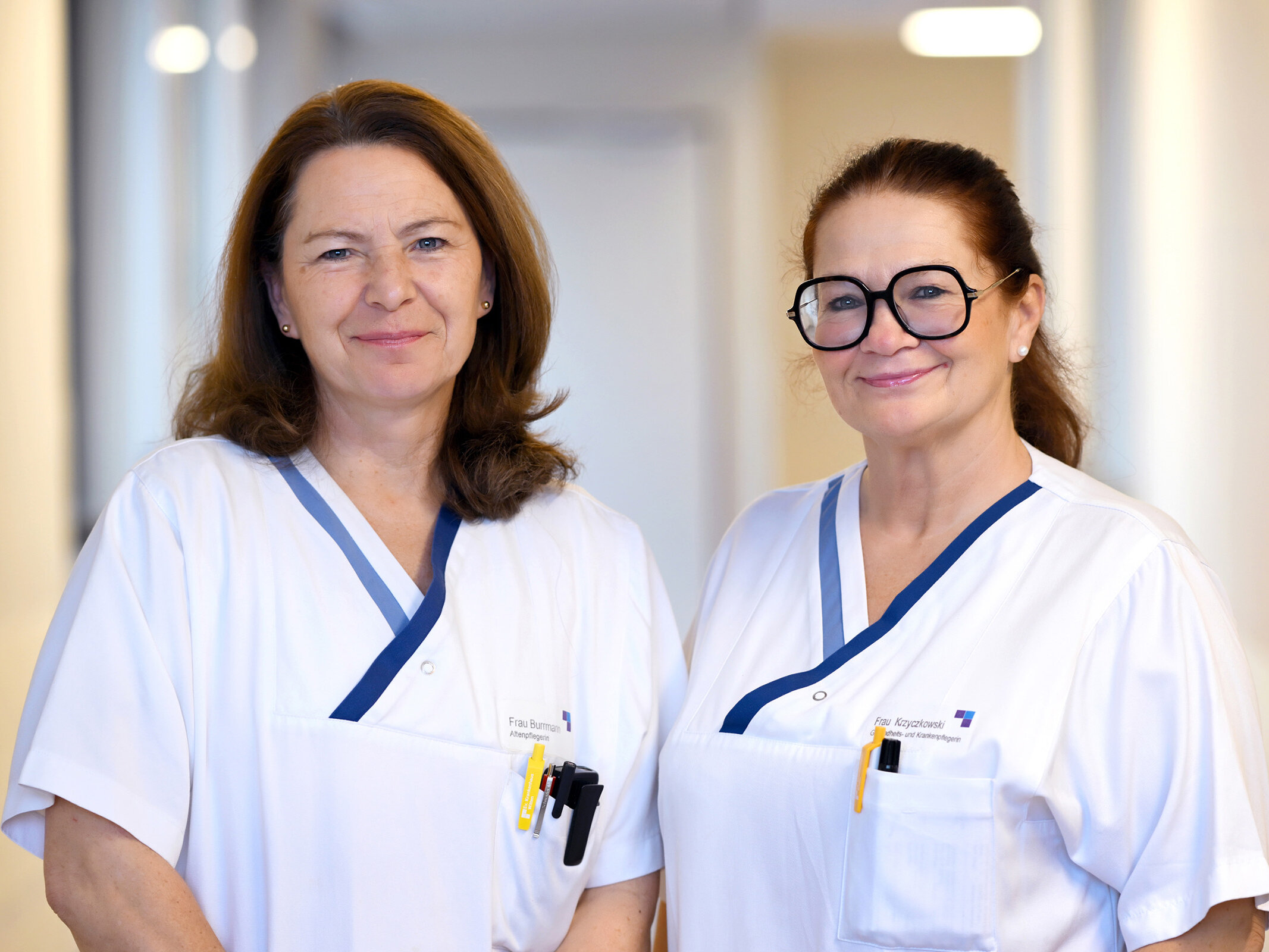 Zwei weibliche Mitarbeiter des Gesundheitswesens in weißen Uniformen stehen lächelnd in einem hell erleuchteten Flur.