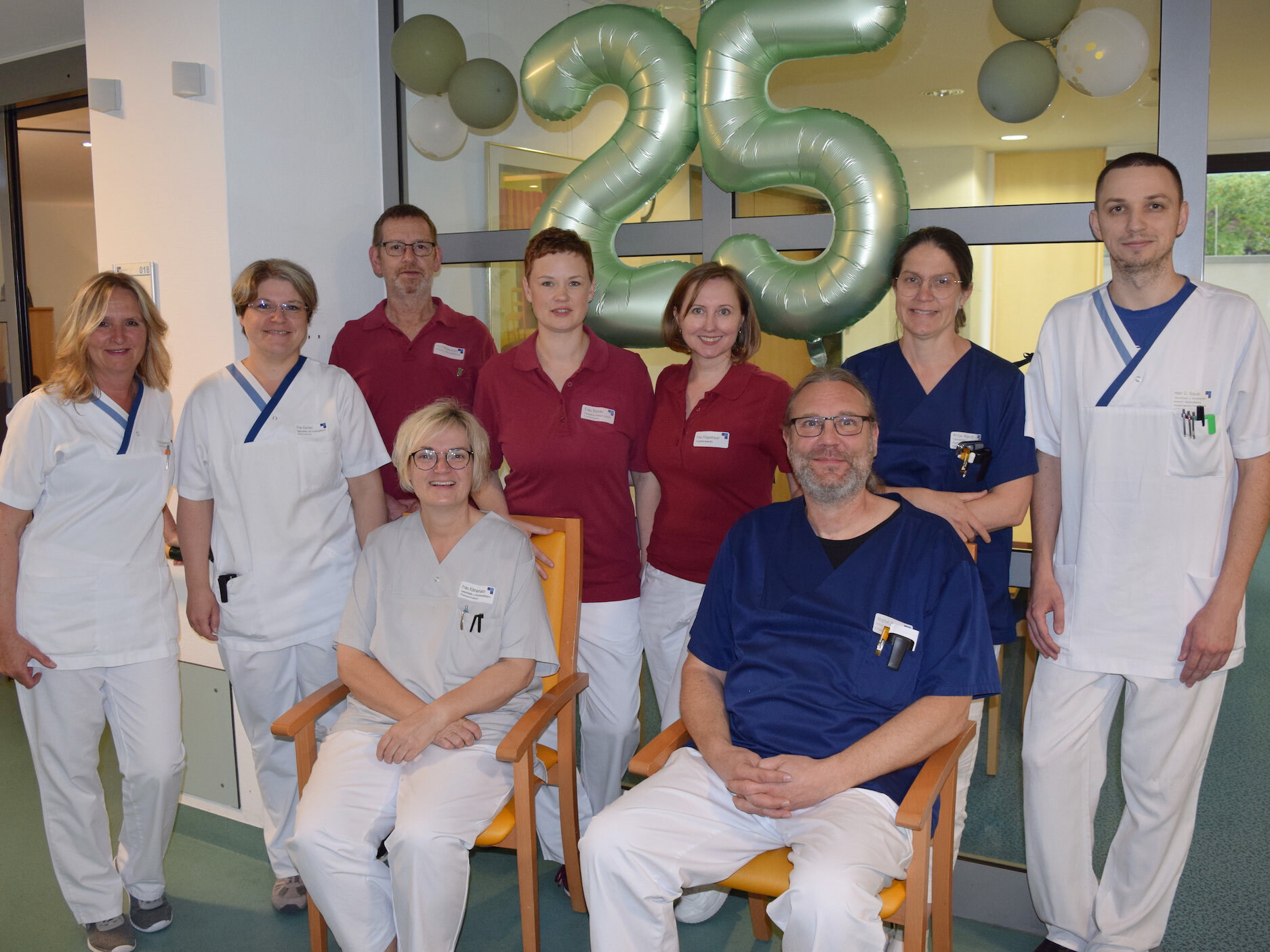 Neun Mitarbeiter des medizinischen Personals posieren in einem Innenraum mit großen "25"-Ballons im Hintergrund, um ein Jubiläum zu feiern.