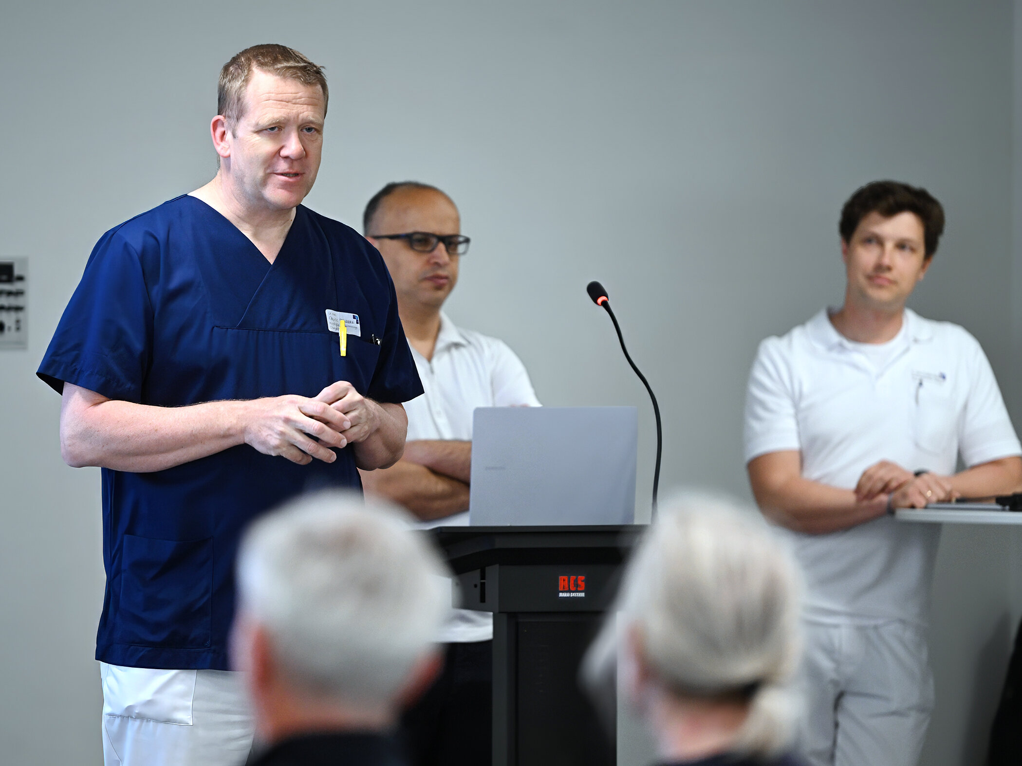 Ein Mann im blauen Kassak spricht vor Menschen. Im Hintergrund stehen zwei Männer an einem Rednerpult in weißen Poloshirts.