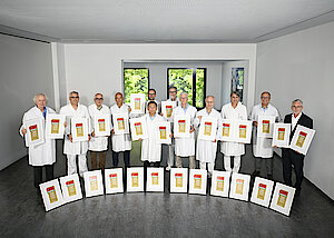 Viele Männer stehen nebeneinander. Bis auf ein Mann tragen alle weiße Kittel. Rechts außen steht ein Mann mit Brille und Bart im dunklen Anzug. Alle halten gerahmte Siegel in den Händen. Weitere Rahmen stehen davor.  den in