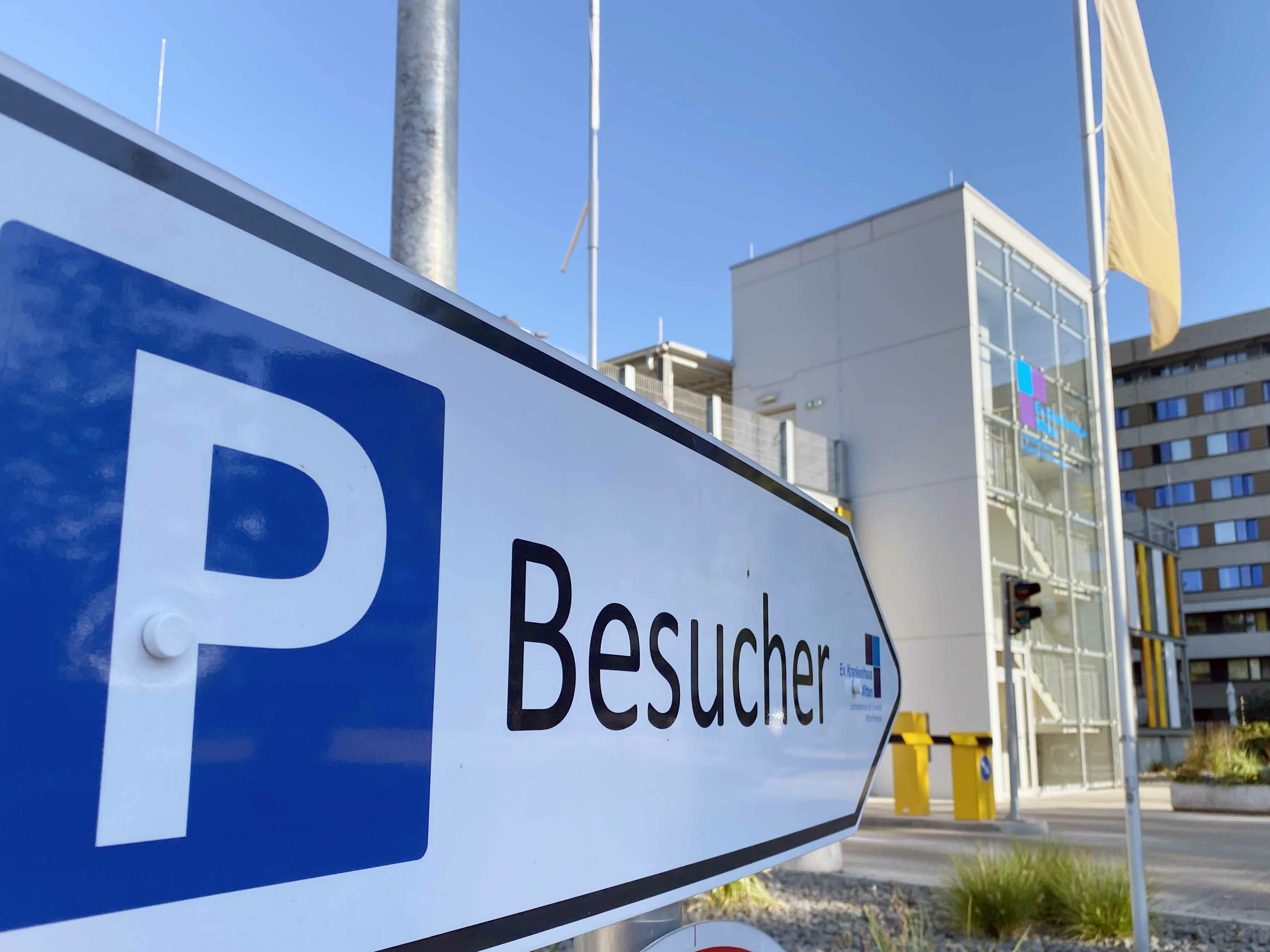 Nahaufnahme eines "Besucher"-Parkplatzschilds in der Nähe eines modernen Gebäudes unter strahlend blauem Himmel.