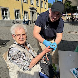 Eine ältere Frau sitzt auf einem Stuhl. Ein Mann in schwarzer Kleidung mit Handschuhen piekst mit einer Nadel in ihren Finger.
