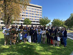 Eine Gruppe von Männern und Frauen in schicker Kleidung steht vor einem hohen Gebäude. Sie halten Blumen in den Händen und lächeln.