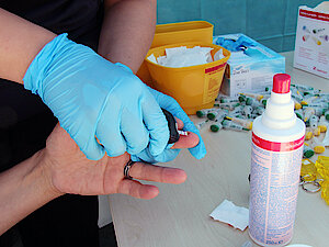 Eine behandschuhte Hand führt einen Blutzuckertest am Finger durch, wobei Desinfektionsmittel und medizinisches Material auf einem Tisch liegen.