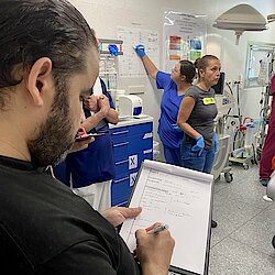 Ein Mann mit schwarzen Haaren und schwarzem T-Shirt notiert etwas auf einem Klemmbrett. Im Hintergrund ist eine sprechende Frau zu sehen. Eine andere notiert etwas auf einer Folie, die an der Wand hängt.