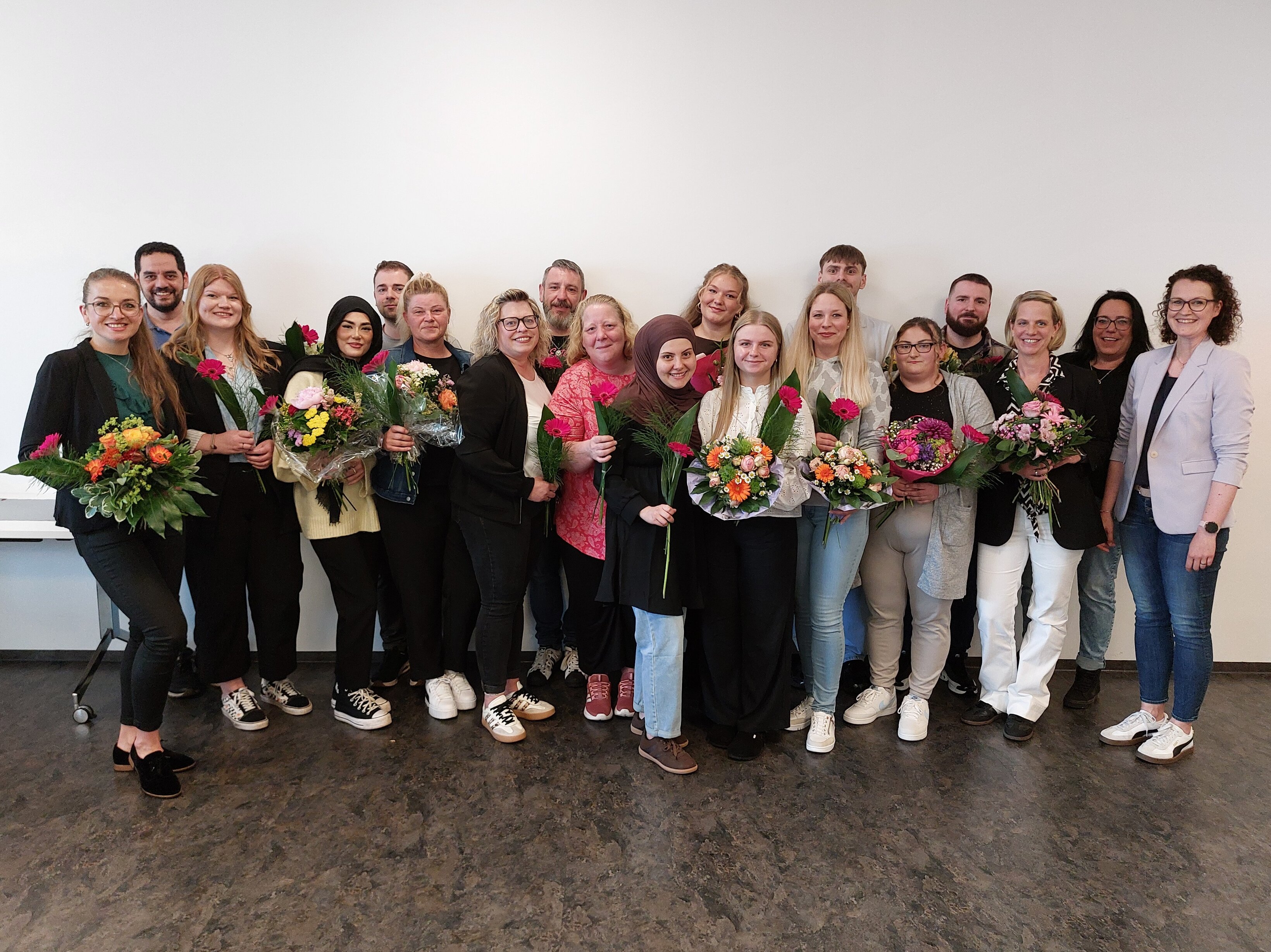 Junge Menschen, teilweise mit Blumen in den Händen, stehen in mehreren Reihen nebeneinander vor einer weißen Wand und lächeln.