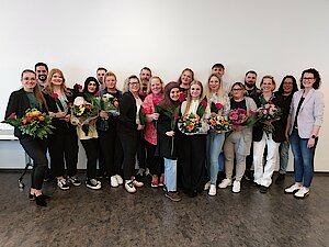Junge Menschen, teilweise mit Blumen in den Händen, stehen in mehreren Reihen nebeneinander vor einer weißen Wand und lächeln.