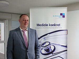 Ein Mann in einem hellen Anzug steht neben einem Plakat mit der Aufschrift Medizin konkret und dem Logo des EvK Witten