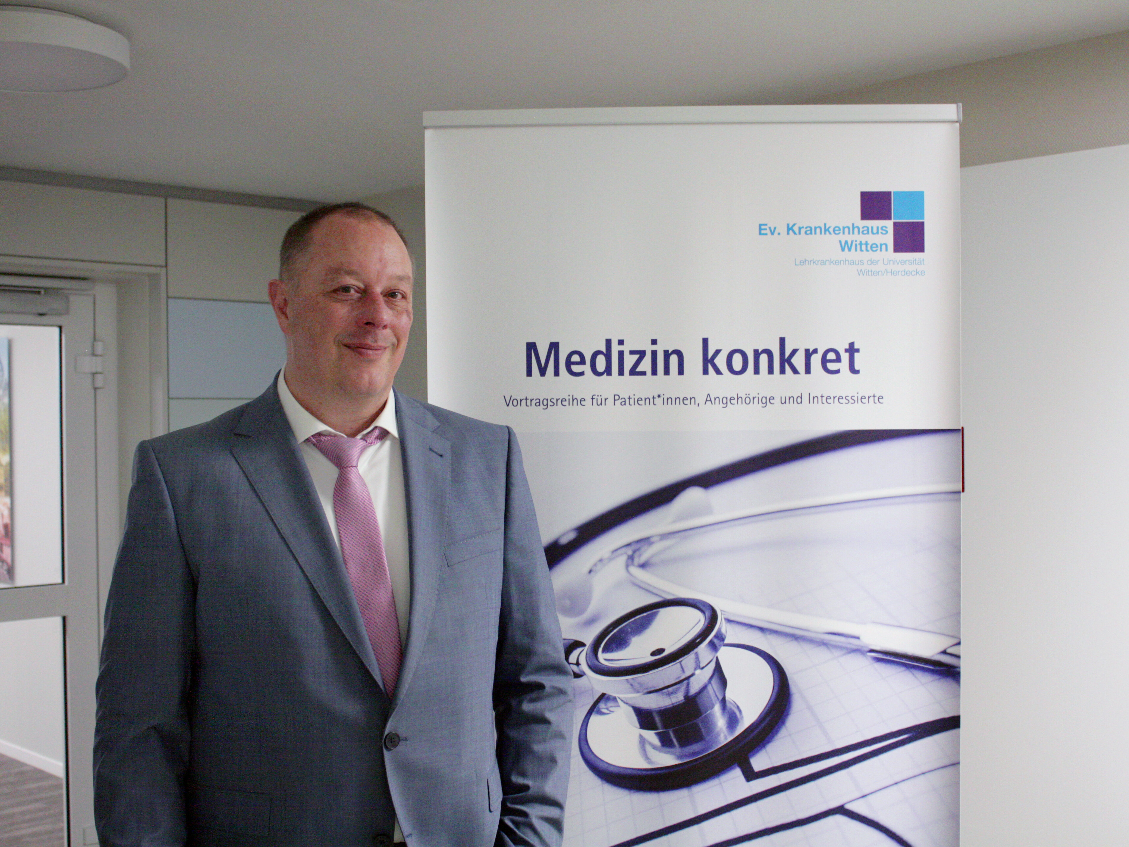 Ein Mann in einem hellen Anzug steht neben einem Plakat mit der Aufschrift Medizin konkret und dem Logo des EvK Witten