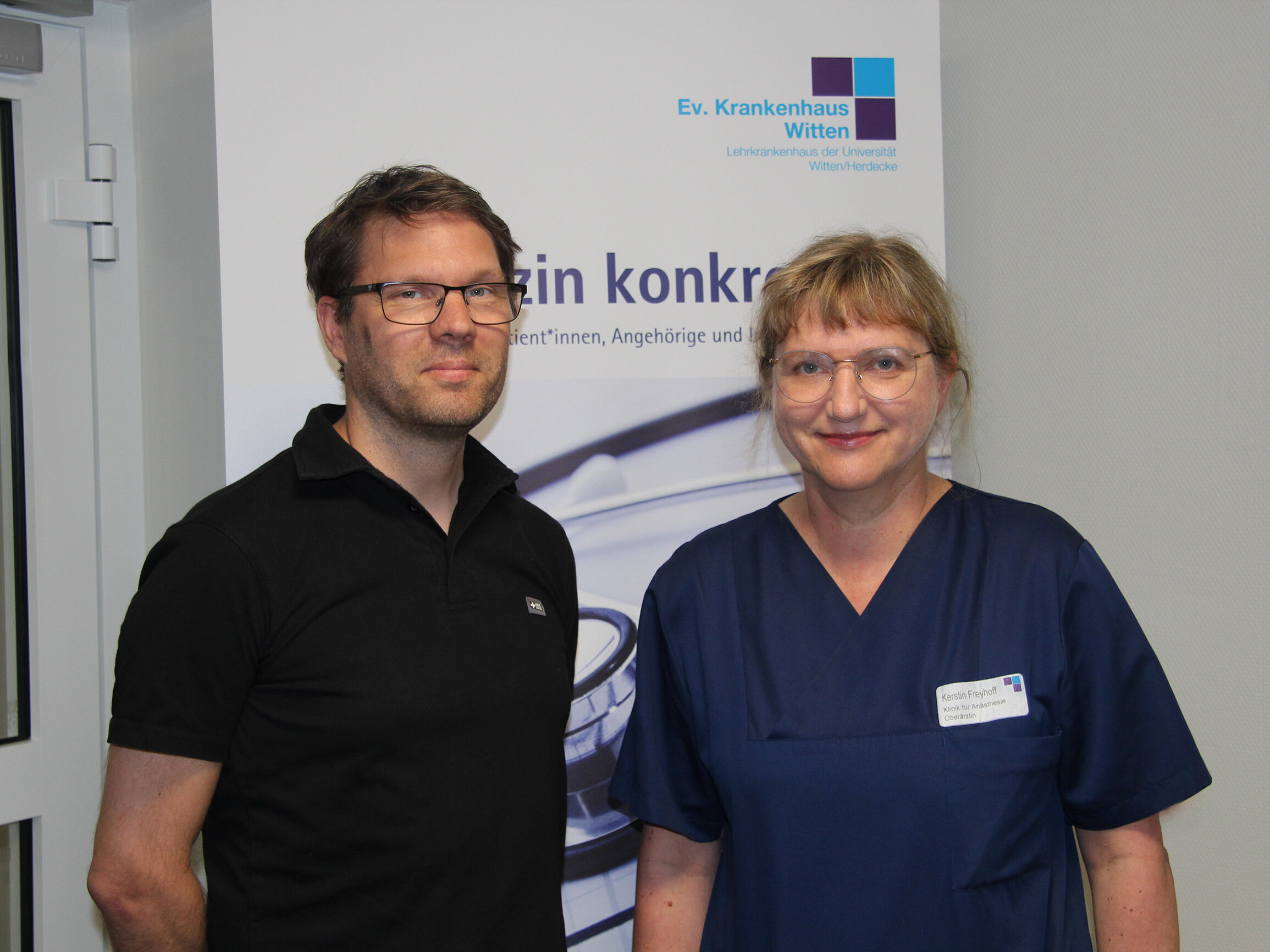Ein Mann in schwarzem Poloshirt und eine Frau in blauer Arztkleidung stehen vor einem Aufsteller mit der Aufschrift Medizin konkret.