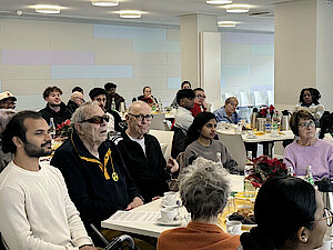 Junge und alte Menschen sitzen in einem Raum an Tischen. Auf den Tischen stehen Getränke und weihnachtliches Gebäck.