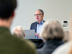 Mann mit Sakko, Hemd und Brille steht am Rednerpult und erzählt. Im Hintergrund sind unscharf Menschen von hinten zu sehen.