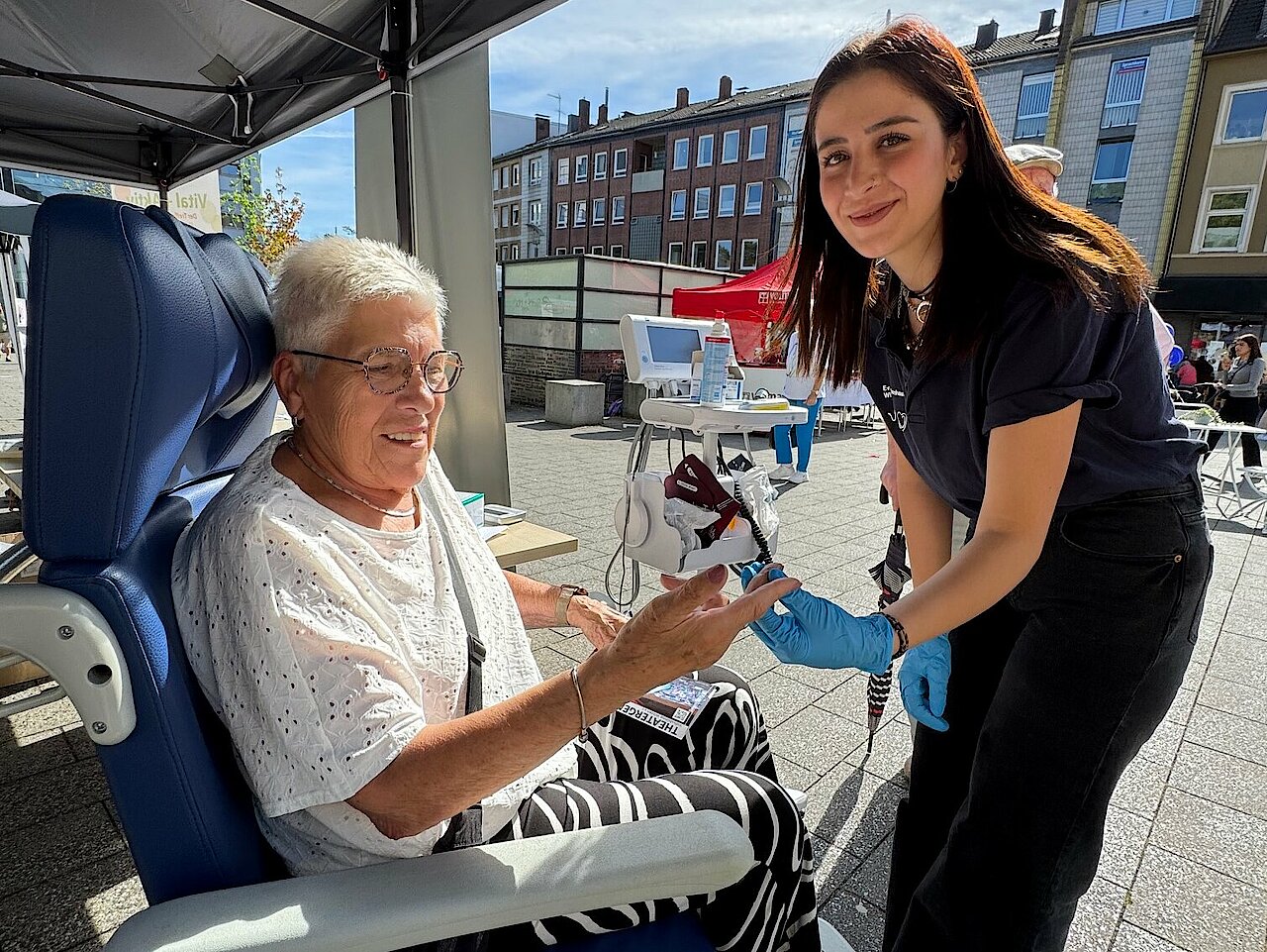 Eine ältere Frau sitzt in einem Rollstuhl. Eine dunkelhaarige jüngere Frau misst den Blutzucker. 