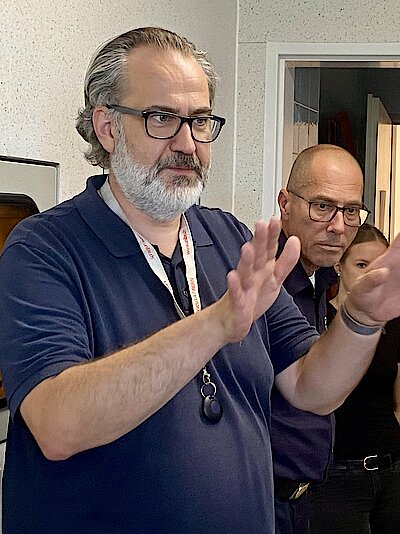 Ein Mann in blauem Polo-Shirt steht vor einer Wand, gestikuliert mit den Händen und spricht. Mehrere Menschen stehen um ihn herum und hören zu. 