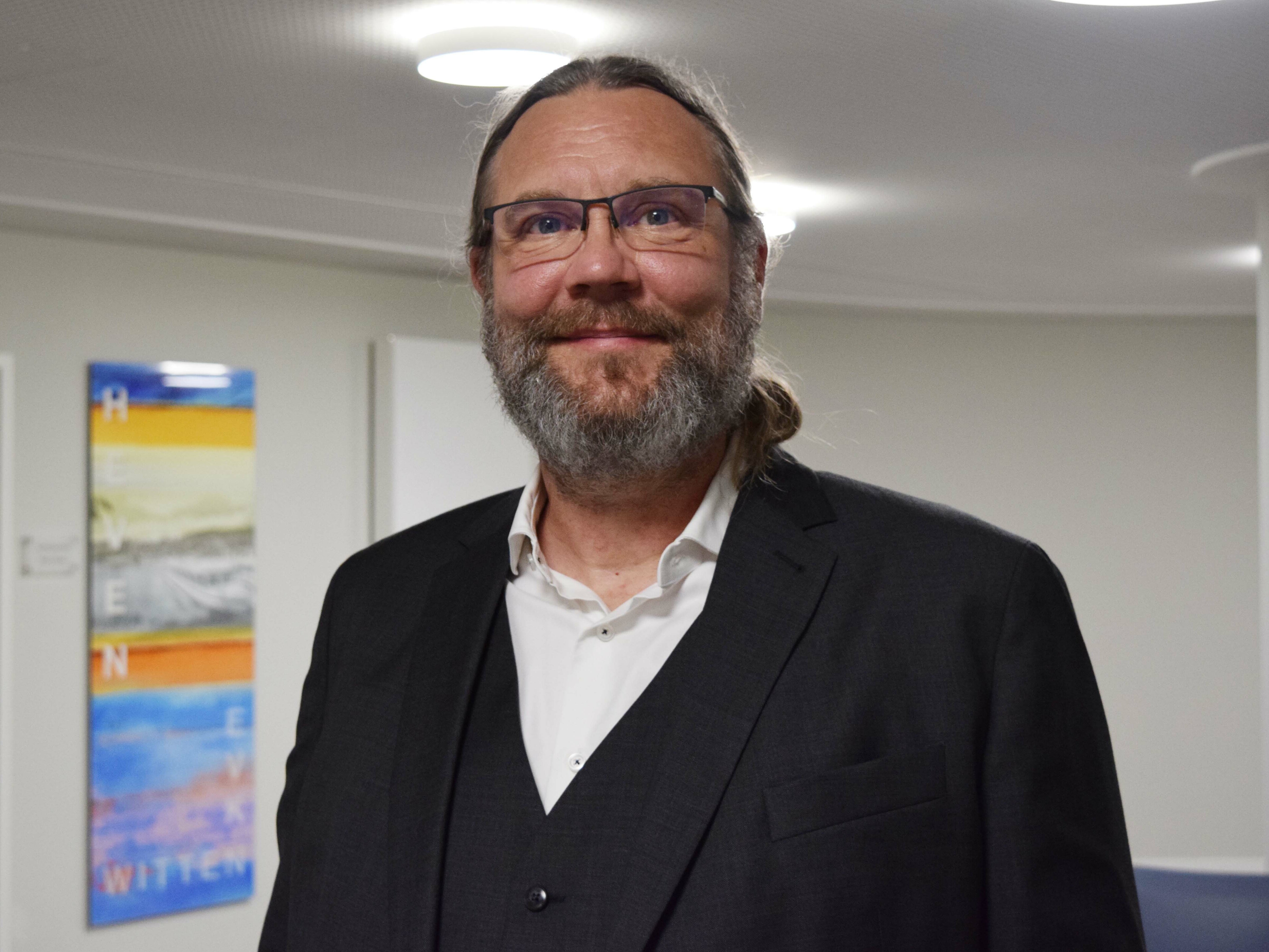 Ein Mann mit Brille, Bart und Anzug lächelt in einem Raum, in dem bunte Kunstwerke an der Wand hinter ihm hängen.