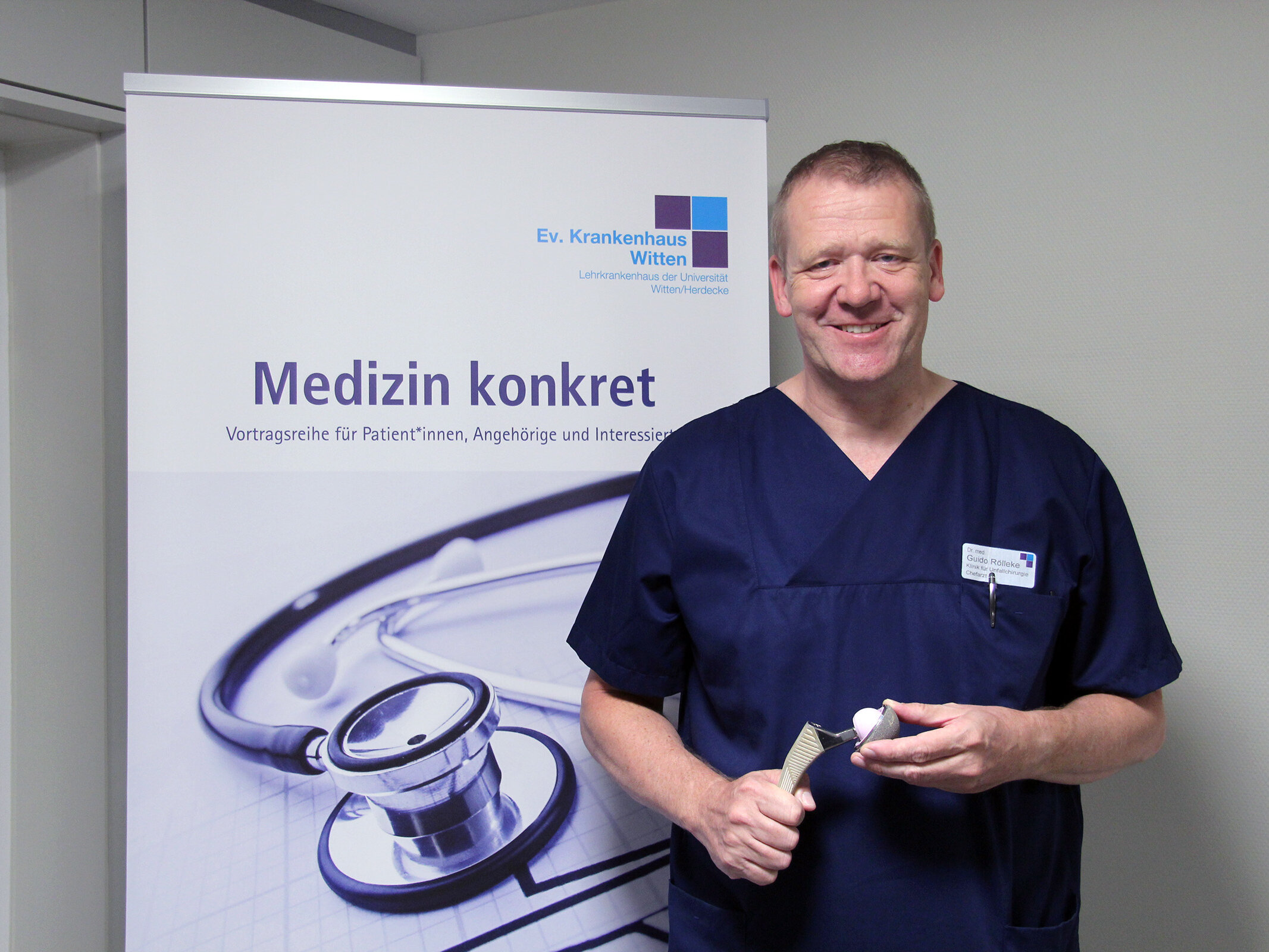 Ein Arzt steht mit einem künstlichen Kniegelenk vor einem Banner mit der Aufschrift Medizin konkret.