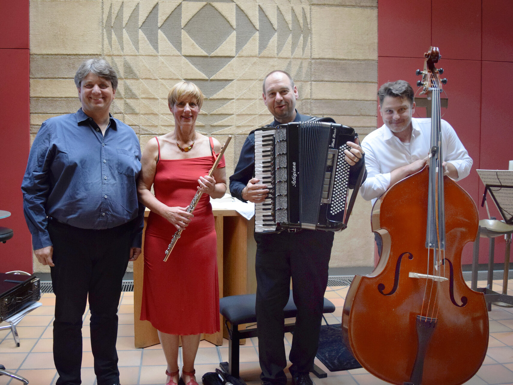 Vier Musiker posieren mit ihren Instrumenten: Klavier, Flöte, Akkordeon und Kontrabass, vor einem gemusterten Bühnenhintergrund.