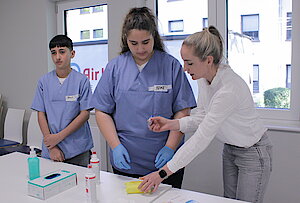 Eine Frau in hält eine Spritze in der Hand und erklärt einem Jungen und einem Mädchen etwas. 