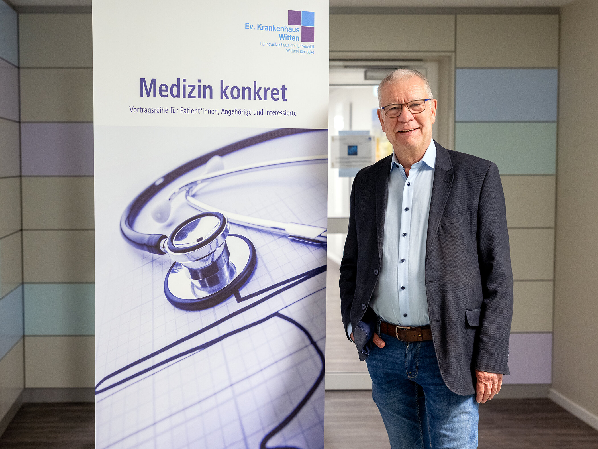 Ein Mann in Hemd und Sakko steht vor einem Roll-Up. Darauf steht "Medizin konkret".