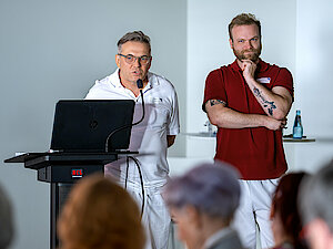 Zwei Männer stehen vor einem Rednerpult. Der Mann in weißer Kleidung spricht. Der Mann daneben trägt eine weiße Hose und ein rotes Polo-Shirt und hört zu.