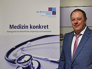 Ein Mann in einem blauen Anzug steht neben einem Plakat für einen medizinischen Vortrag mit Stethoskop und deutschem Text.