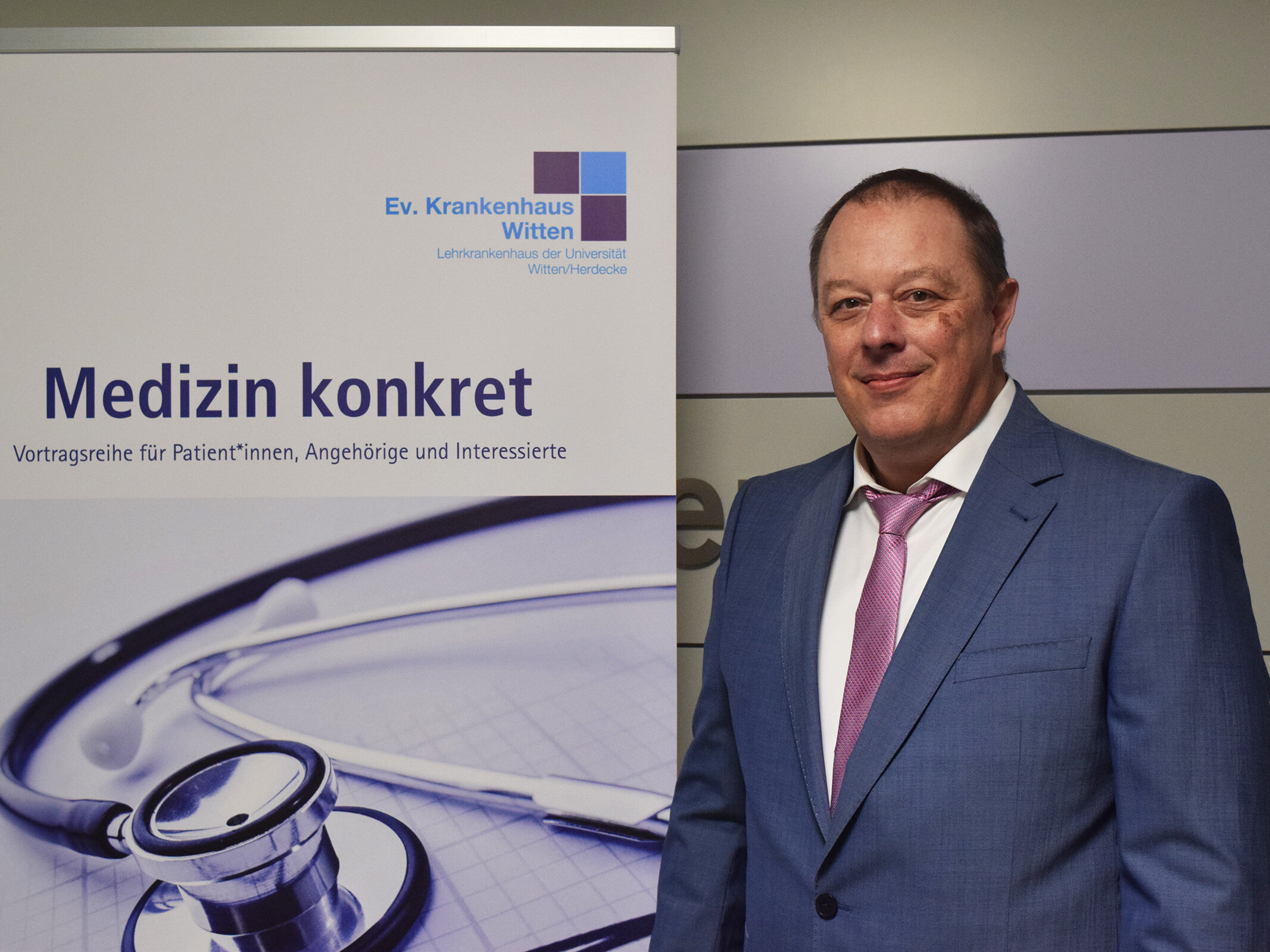Ein Mann in einem blauen Anzug steht neben einem Plakat für einen medizinischen Vortrag mit Stethoskop und deutschem Text.