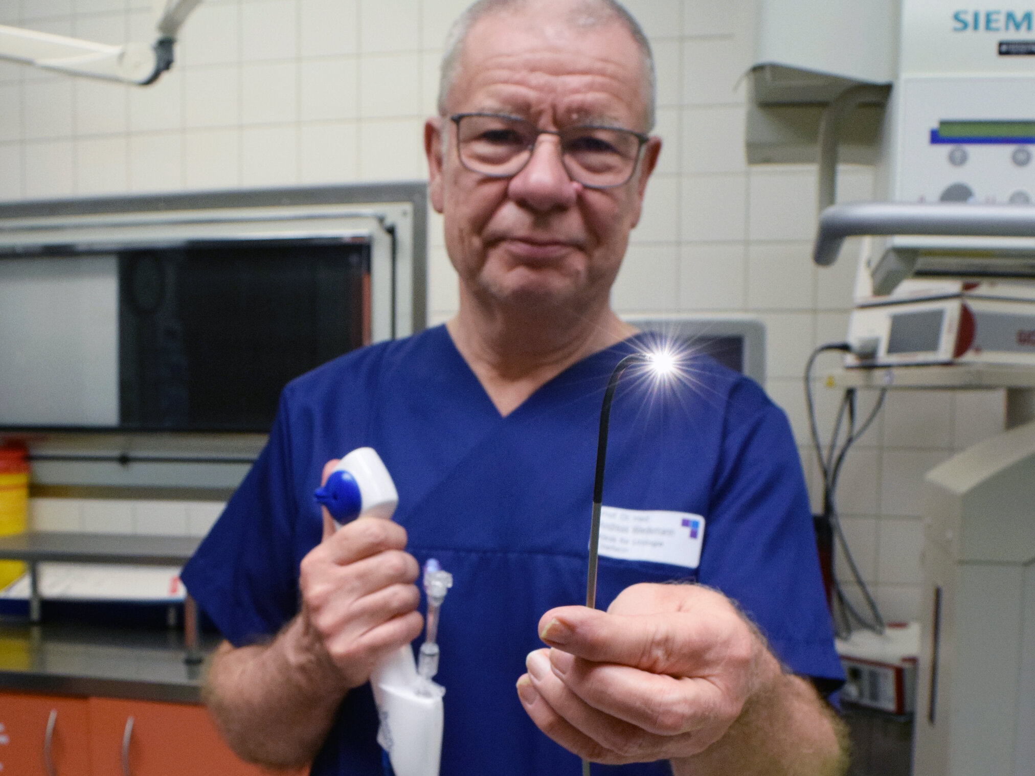 Älterer Mann in OP-Kleidung hält ein medizinisches Gerät mit einer Lampe in  einem Untersuchungsraum.