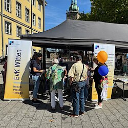 Ein grauer Pavillon steht neben einem gelben Gebäude. Davor stehen Menschen. Auf Bannern steht EvK Witten geschrieben.