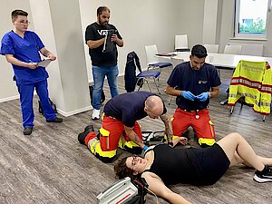 Zwei Rettungssanitäter knien auf dem Boden und behandeln eine auf dem Boden liegende Frau. Im Hintergrund stehen eine Mann und eine Frau und beobachten die Szene.