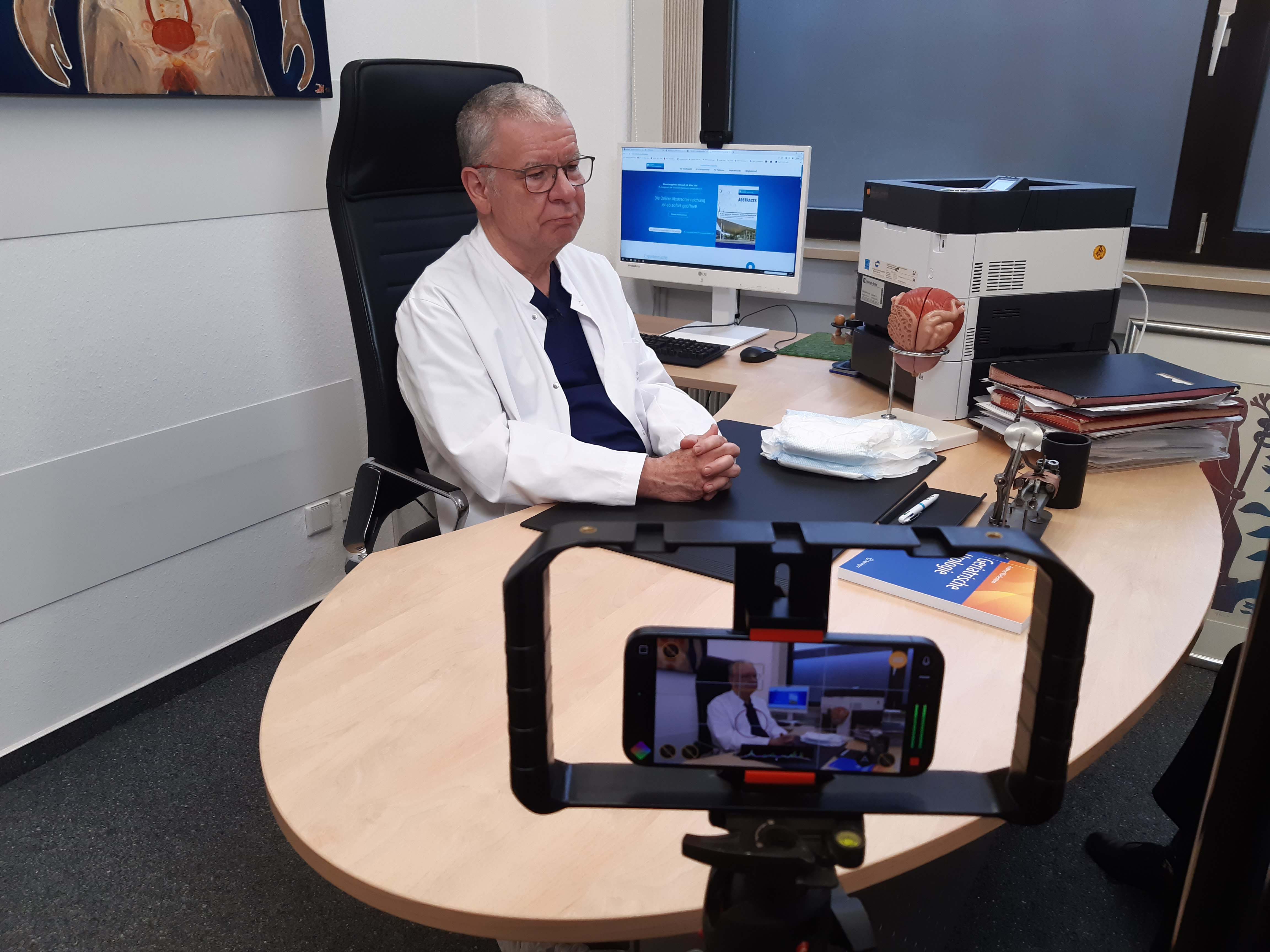 Ein Arzt sitzt an seinem Schreibtisch und wird von einer Smartphone-Kamera in einem Büro mit medizinischen Akten und einem Computer aufgenommen.
